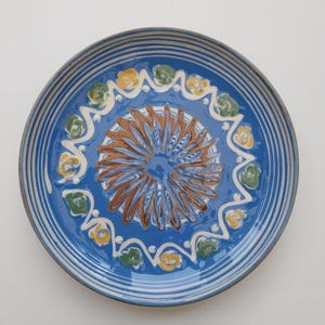 May include: A blue ceramic plate with a white rim and a decorative design in the center. The design features a brown swirl with yellow and green accents.