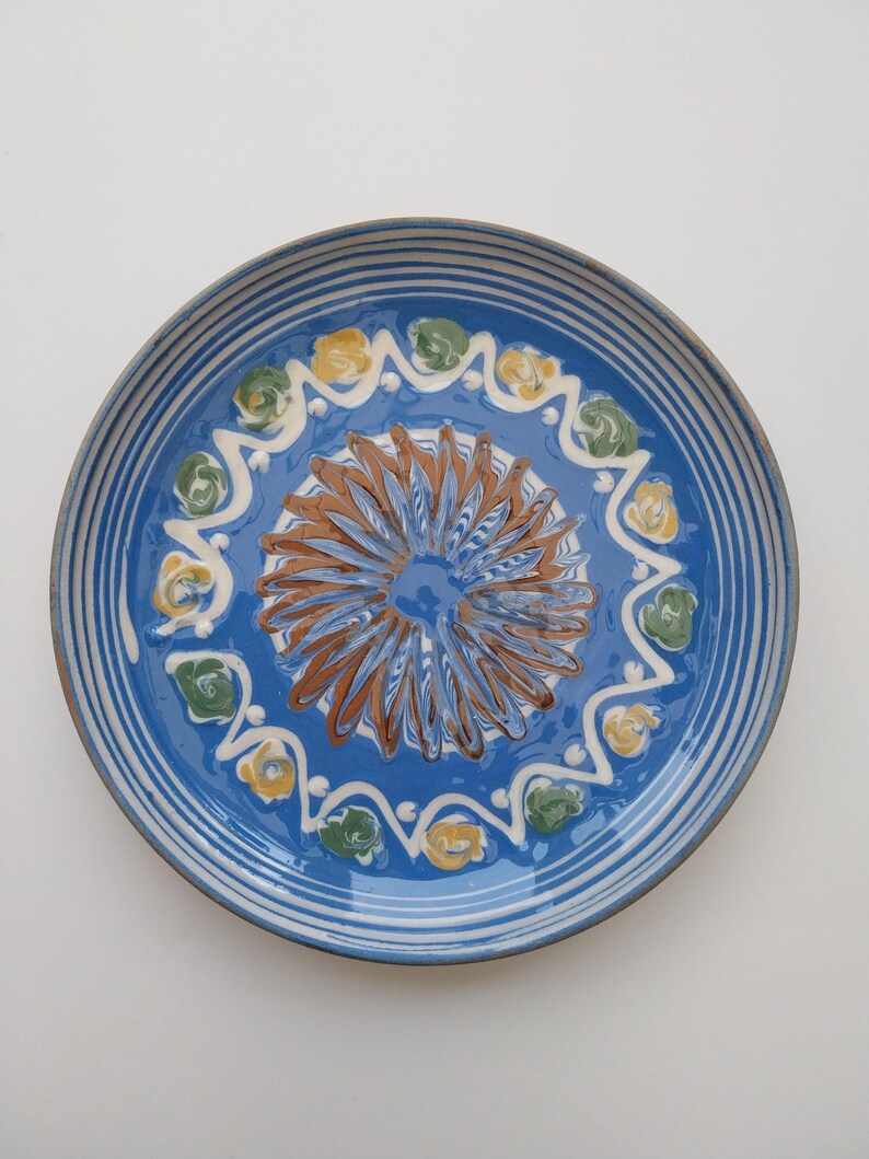 May include: A blue ceramic plate with a white rim and a decorative design in the center. The design features a brown swirl with yellow and green accents.