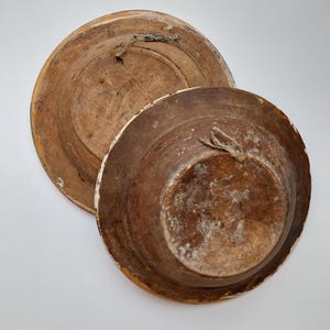 May include: Two antique wooden plates, one stacked on top of the other. The plates are brown with white trim and show signs of wear. Each plate has a loop for hanging.