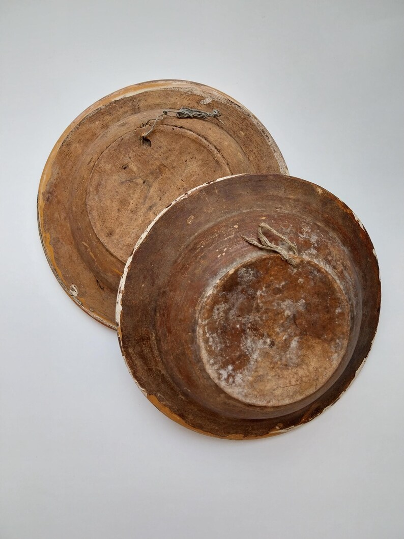 May include: Two antique wooden plates, one stacked on top of the other. The plates are brown with white trim and show signs of wear. Each plate has a loop for hanging.