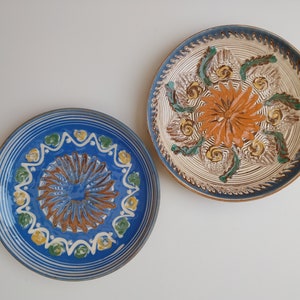 May include: Two decorative ceramic plates with blue and white borders and intricate floral designs in the center. The plate on the left has a blue center with brown and white details, while the plate on the right has a white center with orange and green details.