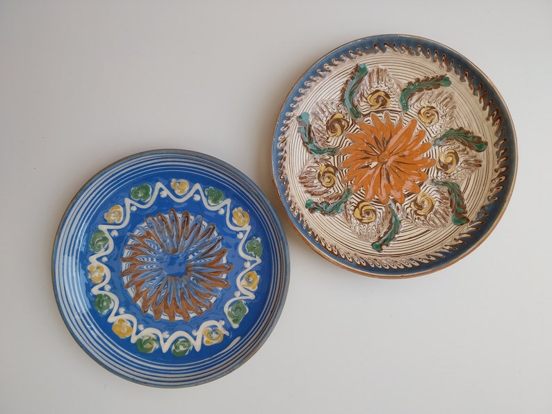 May include: Two decorative ceramic plates with blue and white borders and intricate floral designs in the center. The plate on the left has a blue center with brown and white details, while the plate on the right has a white center with orange and green details.