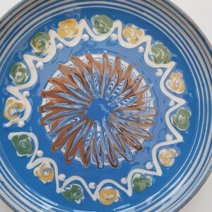 May include: A blue ceramic plate with a white rim and a decorative design in the center. The design features a brown swirl with yellow and green accents.