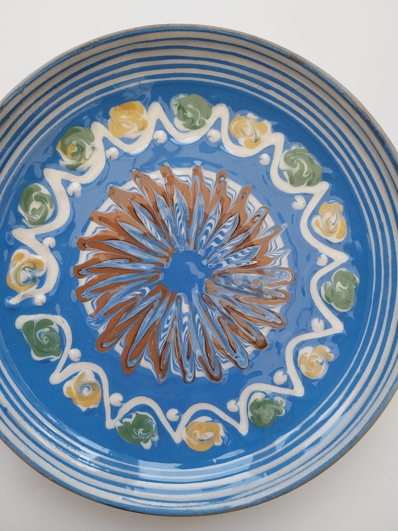 May include: A blue ceramic plate with a white rim and a decorative design in the center. The design features a brown swirl with yellow and green accents.