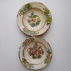 May include: Two antique ceramic plates with hand-painted floral designs. The plates have a cream-coloured base with colourful flowers in the centre and around the rim. The rims have a rustic, aged appearance, adding to their vintage charm.