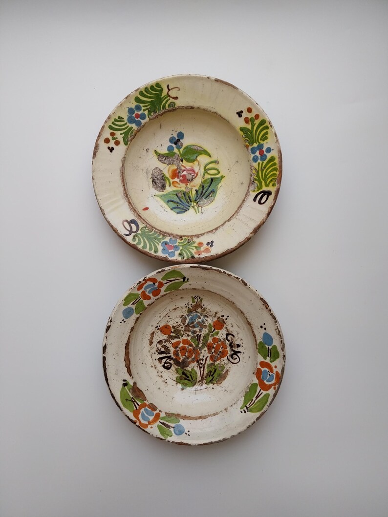 May include: Two antique ceramic plates with hand-painted floral designs. The plates have a cream-coloured base with colourful flowers in the centre and around the rim. The rims have a rustic, aged appearance, adding to their vintage charm.