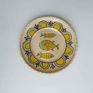 May include: A round, cream-colored ceramic plate with a scalloped edge. The plate features a central design of three yellow and green fish. A decorative border of yellow and green floral shapes surrounds the fish.