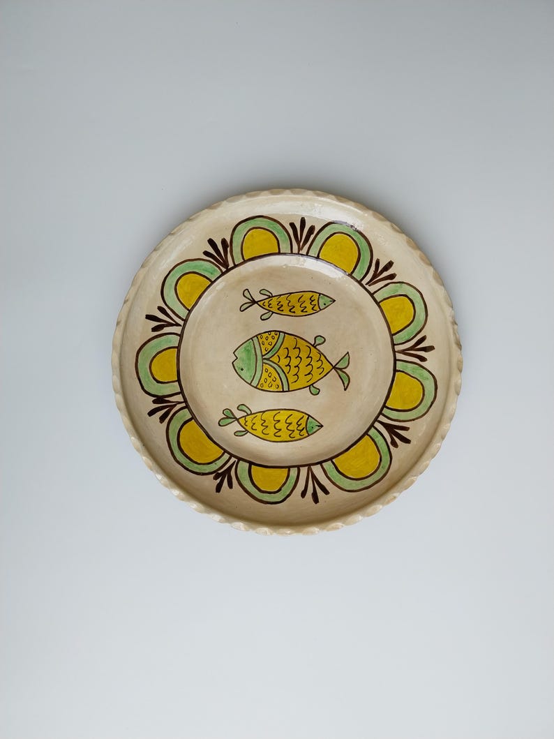 May include: A round, cream-colored ceramic plate with a scalloped edge. The plate features a central design of three yellow and green fish. A decorative border of yellow and green floral shapes surrounds the fish.
