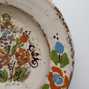 May include: Off-white ceramic plate with hand-painted floral design in orange, blue, and green. The plate has a distressed, rustic look with a worn, brown rim and visible imperfections. The plate features a floral pattern and a handwritten inscription.
