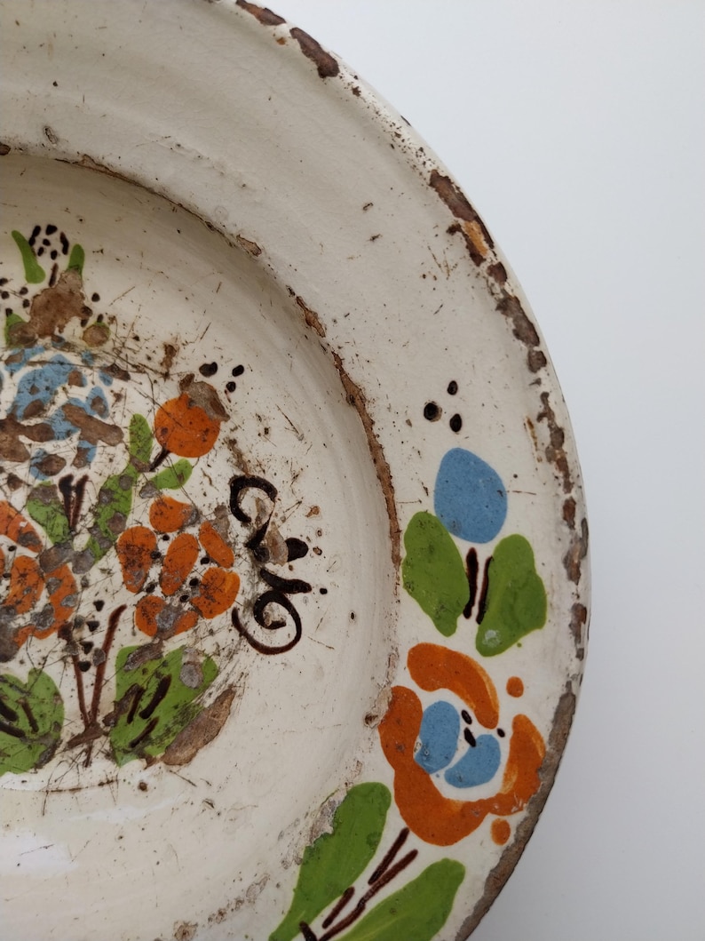 May include: Off-white ceramic plate with hand-painted floral design in orange, blue, and green. The plate has a distressed, rustic look with a worn, brown rim and visible imperfections. The plate features a floral pattern and a handwritten inscription.