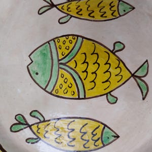 May include: A ceramic plate with a hand-painted design of three yellow fish with green and black accents. The fish are swimming on a cream-colored background. The plate's rim has a yellow and brown border.