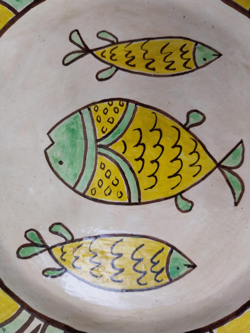 May include: A ceramic plate with a hand-painted design of three yellow fish with green and black accents. The fish are swimming on a cream-colored background. The plate's rim has a yellow and brown border.