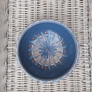 May include: A blue ceramic bowl with a white and yellow floral design and orange dots. The bowl is sitting on a white wicker surface.