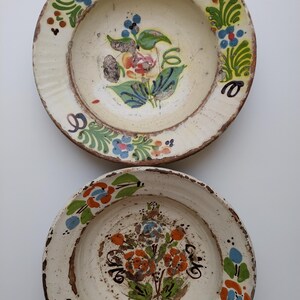 May include: Two antique, hand-painted ceramic plates. The plates have a cream-coloured base with floral designs in shades of blue, orange, green, and red. The rims are a darker brown, and the plates show signs of age and wear.