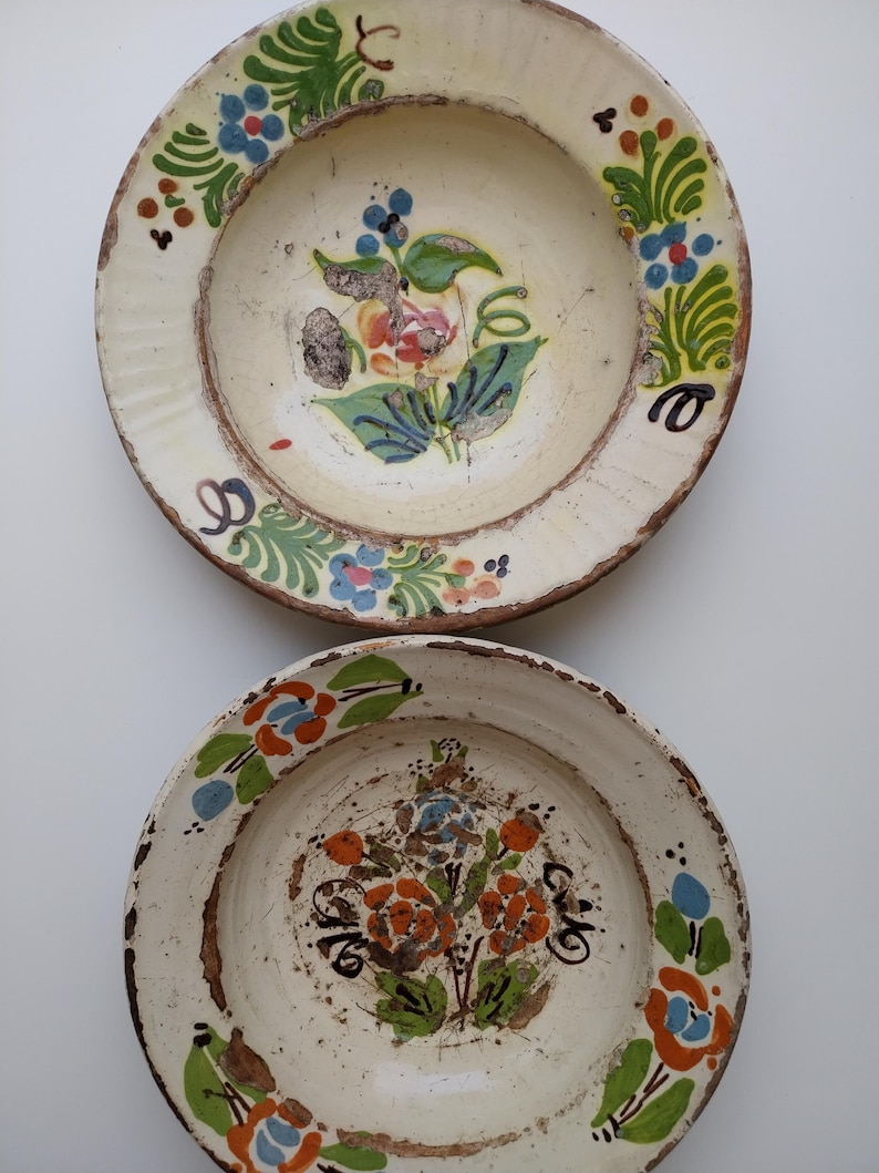May include: Two antique, hand-painted ceramic plates. The plates have a cream-coloured base with floral designs in shades of blue, orange, green, and red. The rims are a darker brown, and the plates show signs of age and wear.