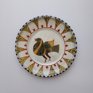 May include: A hand-painted ceramic plate featuring a bird design. The bird is rendered in green, yellow, and blue, with a patterned body and wing. The plate's rim has a repeating pattern of blue, yellow, and red accents, adding to its decorative appeal.