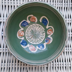 May include: A green ceramic bowl with a white, blue, and orange floral design. The bowl is sitting on a white wicker surface.