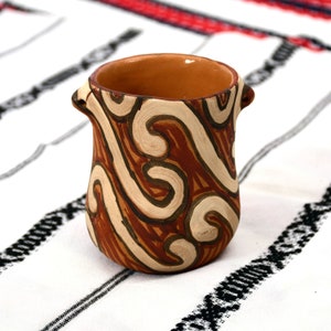 Könnte beinhalten: Eine kleine, braune Keramiktasse mit einem weißen und braunen Wirbelmuster. Die Tasse hat zwei kleine Griffe an den Seiten.