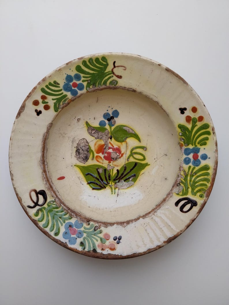 May include: A circular, cream-coloured ceramic plate with a hand-painted floral design. The plate features a central flower with red and orange petals, surrounded by green leaves and blue and pink flowers. The rim has a brown edge and decorative painted accents.