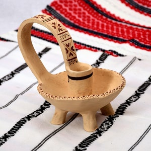May include: A small, beige ceramic candle holder with a handle and three legs. The holder features a brown and red geometric design.