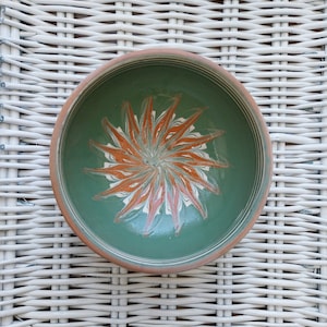 May include: A green ceramic bowl with a hand-painted orange and white floral design. The bowl is on a white wicker surface.