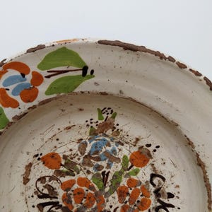 May include: A white ceramic plate with a hand-painted floral design. The flowers are orange, blue, and green, with brown and black accents. The rim of the plate has a rustic, textured brown edge.