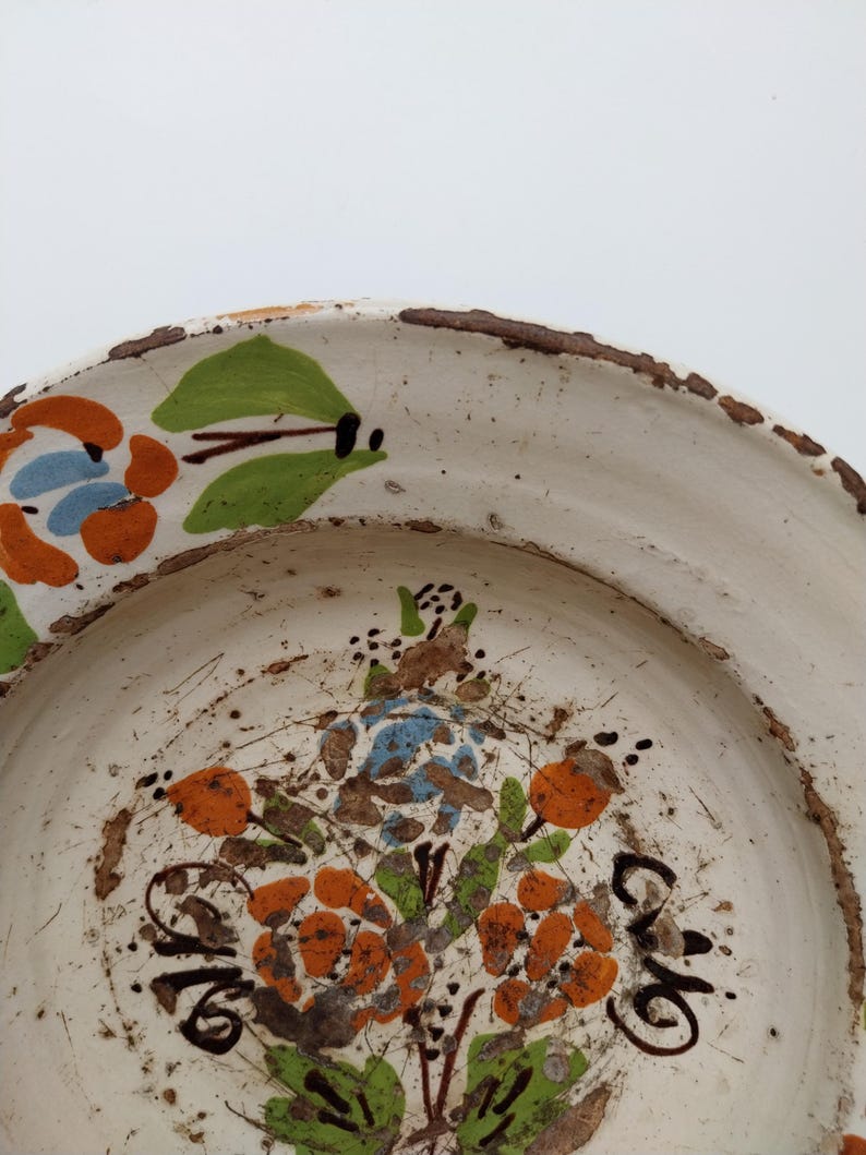May include: A white ceramic plate with a hand-painted floral design. The flowers are orange, blue, and green, with brown and black accents. The rim of the plate has a rustic, textured brown edge.