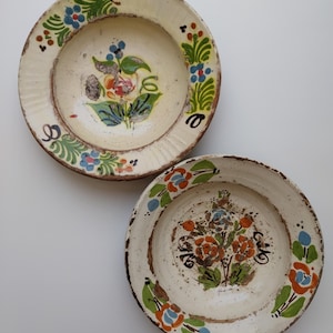 May include: Two antique ceramic plates with floral designs. The plates have a cream-coloured base with hand-painted flowers in shades of orange, blue, and green. The rims are textured and the plates have a rustic, aged appearance.