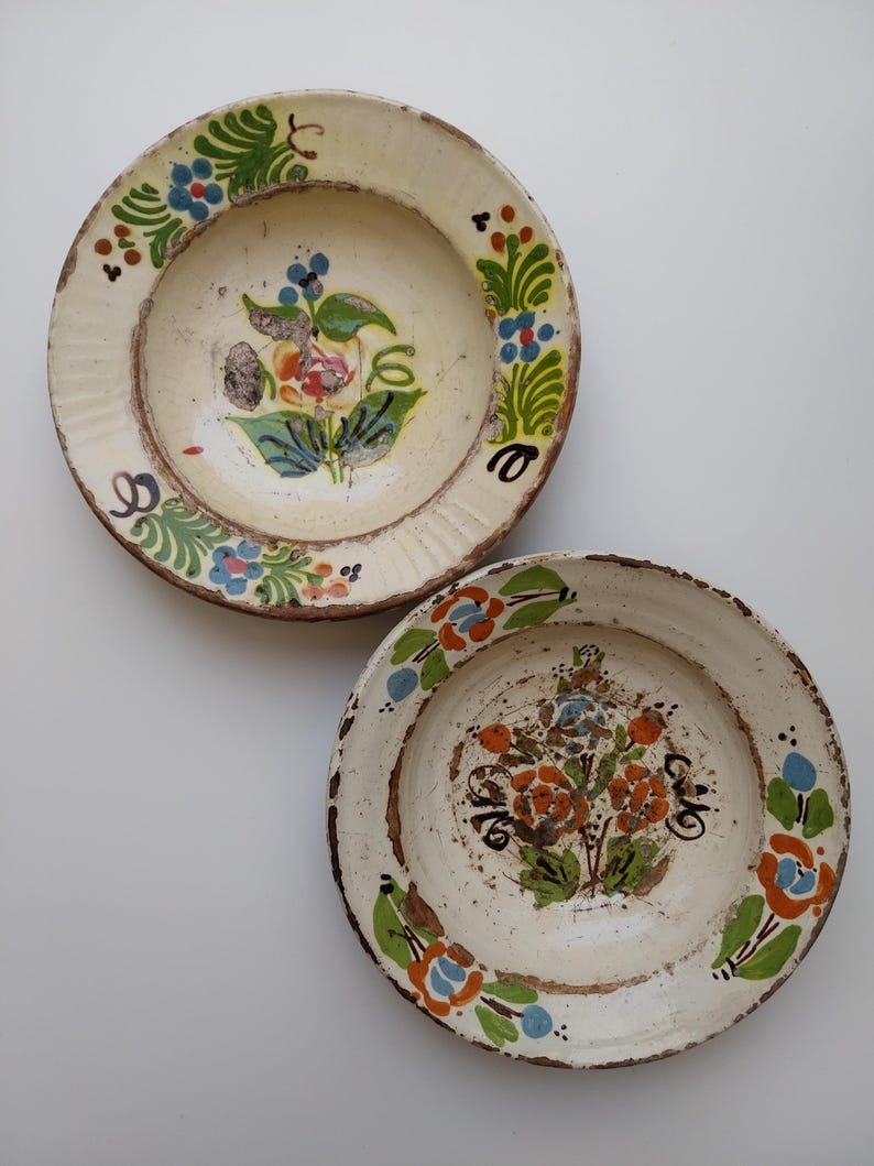 May include: Two antique ceramic plates with floral designs. The plates have a cream-coloured base with hand-painted flowers in shades of orange, blue, and green. The rims are textured and the plates have a rustic, aged appearance.