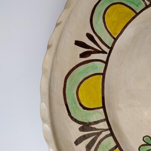 May include: A handmade ceramic plate with a scalloped edge. The plate features a cream-colored base with a painted design of green, yellow, and brown floral elements. The design is symmetrical and appears to be hand-painted.