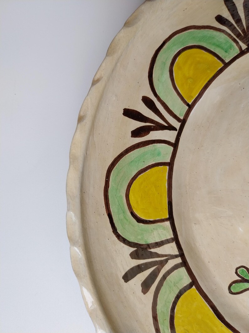 May include: A handmade ceramic plate with a scalloped edge. The plate features a cream-colored base with a painted design of green, yellow, and brown floral elements. The design is symmetrical and appears to be hand-painted.