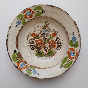 May include: A white ceramic plate featuring a hand-painted floral design. The flowers are orange and blue with green leaves. The rim is brown and the plate has a rustic, aged appearance.