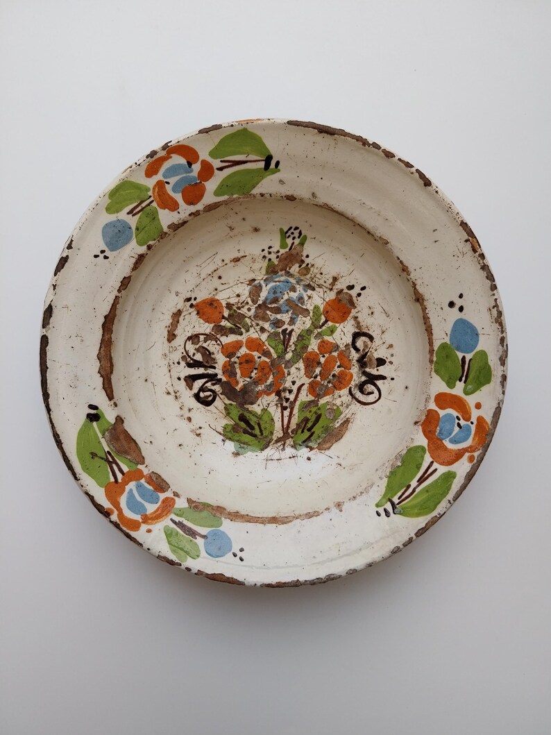 May include: A white ceramic plate featuring a hand-painted floral design. The flowers are orange and blue with green leaves. The rim is brown and the plate has a rustic, aged appearance.