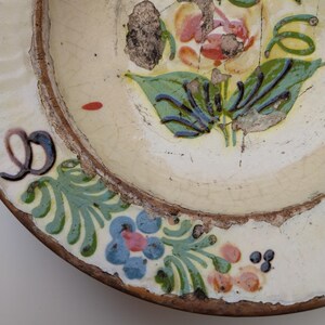 May include: An antique ceramic plate with a floral pattern. The plate has an off-white base, a brown rim, and is adorned with blue, pink, and green floral motifs. The design shows signs of age and wear, enhancing its vintage appeal.