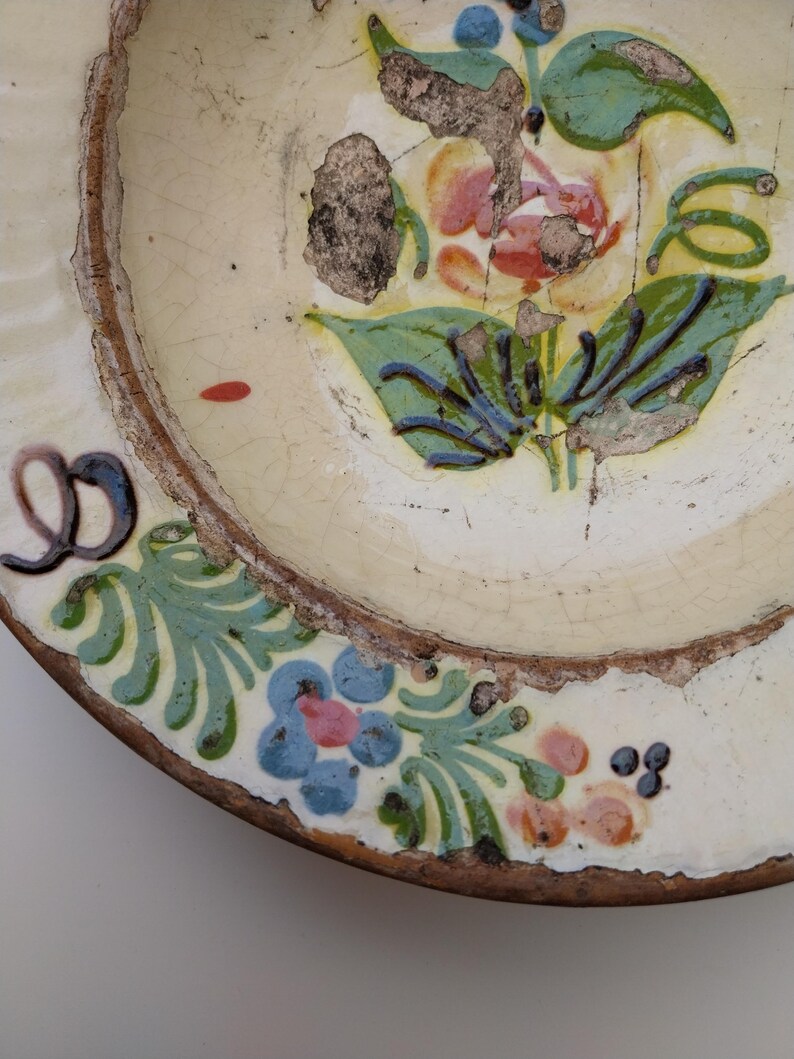 May include: An antique ceramic plate with a floral pattern. The plate has an off-white base, a brown rim, and is adorned with blue, pink, and green floral motifs. The design shows signs of age and wear, enhancing its vintage appeal.