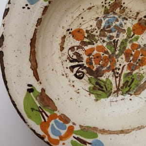 May include: Cream-coloured ceramic plate with a distressed finish. It features hand-painted floral patterns in orange, blue, and green, with brown detailing. The rim has a worn, brown edge, enhancing its vintage appeal. The plate has a rustic look.