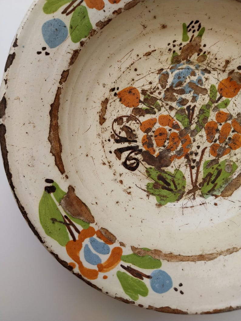 May include: Cream-coloured ceramic plate with a distressed finish. It features hand-painted floral patterns in orange, blue, and green, with brown detailing. The rim has a worn, brown edge, enhancing its vintage appeal. The plate has a rustic look.