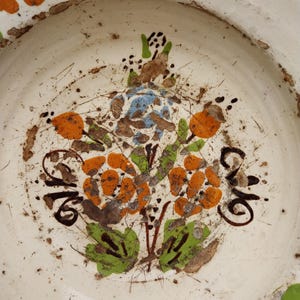 May include: A white ceramic plate with a hand-painted floral design. The flowers are orange, blue, and brown, with green leaves. The plate has a rustic, aged appearance with visible imperfections.