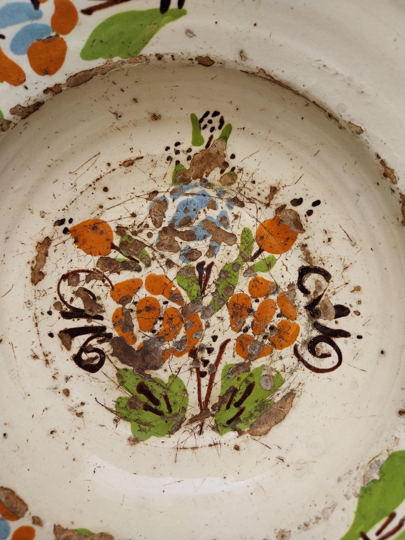 May include: A white ceramic plate with a hand-painted floral design. The flowers are orange, blue, and brown, with green leaves. The plate has a rustic, aged appearance with visible imperfections.