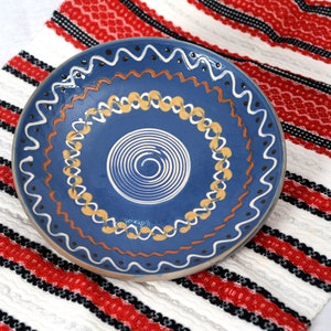 May include: A blue and white ceramic plate with a spiral design in the center. The plate has a decorative border with yellow, orange, and black accents.