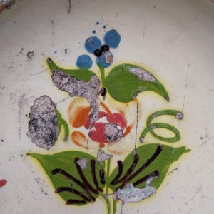May include: Close-up of a ceramic plate with a floral design. The design features a red and orange flower with green leaves and blue berries. The plate has a cream-coloured background with visible wear and tear.