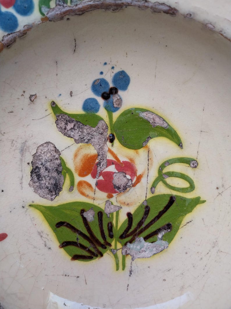 May include: Close-up of a ceramic plate with a floral design. The design features a red and orange flower with green leaves and blue berries. The plate has a cream-coloured background with visible wear and tear.