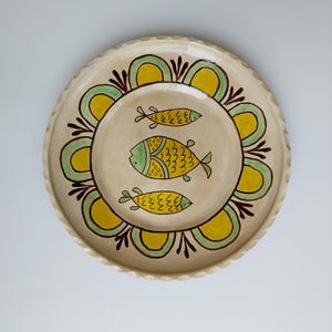May include: A round, cream-colored ceramic plate with a scalloped edge. The plate features a central design of three yellow and green fish. The fish are surrounded by a yellow and green floral pattern with brown accents.