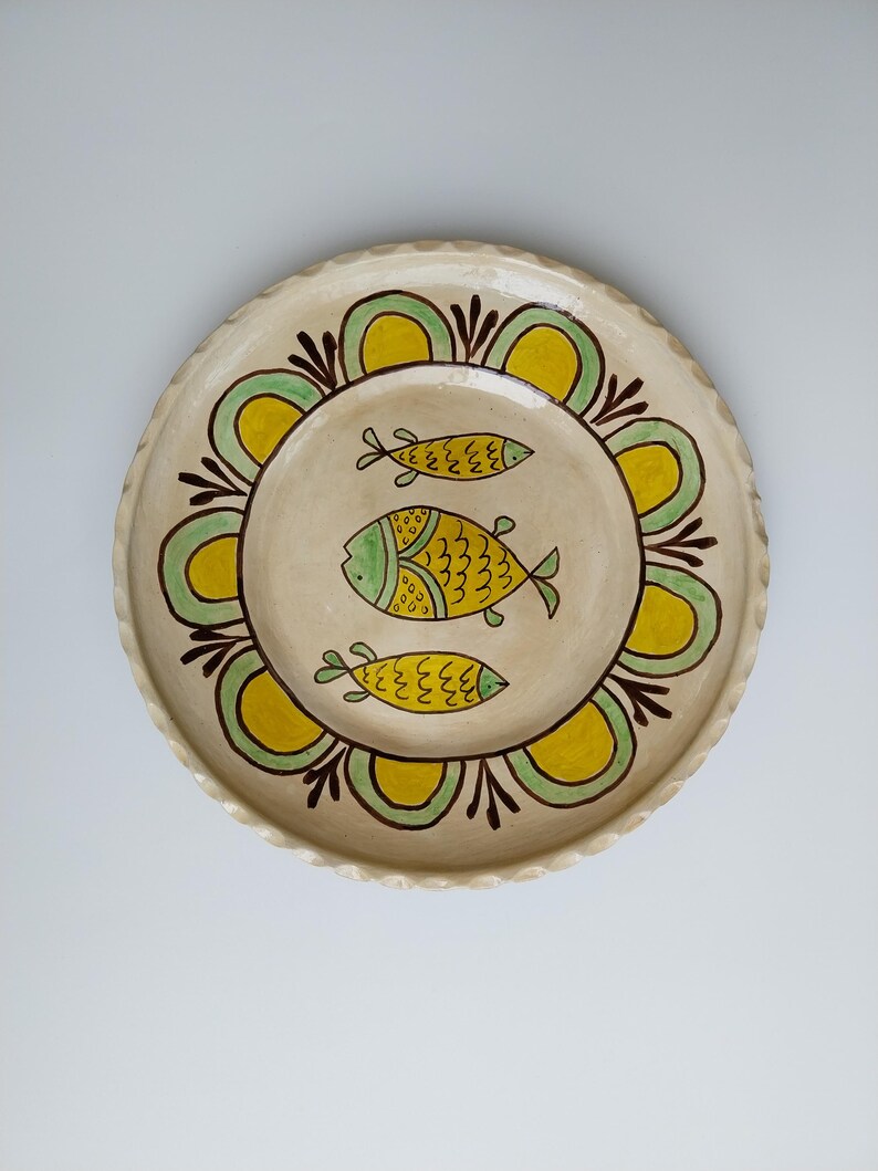 May include: A round, cream-colored ceramic plate with a scalloped edge. The plate features a central design of three yellow and green fish. The fish are surrounded by a yellow and green floral pattern with brown accents.
