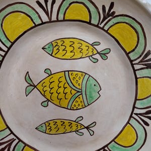 May include: A hand-painted ceramic plate with a cream-colored base. The plate features three yellow fish with green accents in the center. A decorative border of yellow and green arches with brown outlines encircles the fish. The plate has a rustic, folk art style.