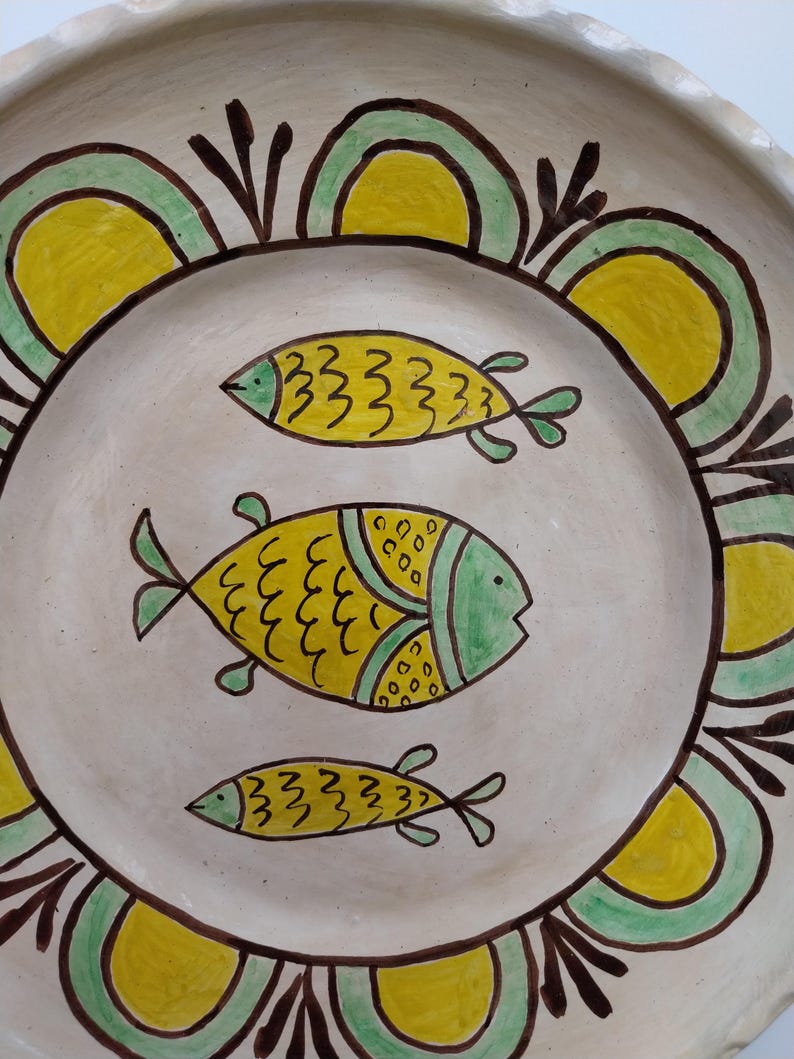 May include: A hand-painted ceramic plate with a cream-colored base. The plate features three yellow fish with green accents in the center. A decorative border of yellow and green arches with brown outlines encircles the fish. The plate has a rustic, folk art style.