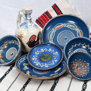 May include: A collection of blue and white ceramic bowls and plates with hand-painted designs. The bowls and plates feature swirling patterns, floral motifs, and geometric shapes. The pottery is displayed on a white and red striped tablecloth.