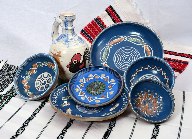 May include: A collection of blue and white ceramic bowls and plates with hand-painted designs. The bowls and plates feature swirling patterns, floral motifs, and geometric shapes. The pottery is displayed on a white and red striped tablecloth.