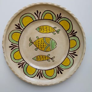 May include: A round, cream-colored ceramic plate with a scalloped edge. The plate features three yellow and green fish designs in the center. A decorative border of yellow and green arches with brown accents surrounds the fish.