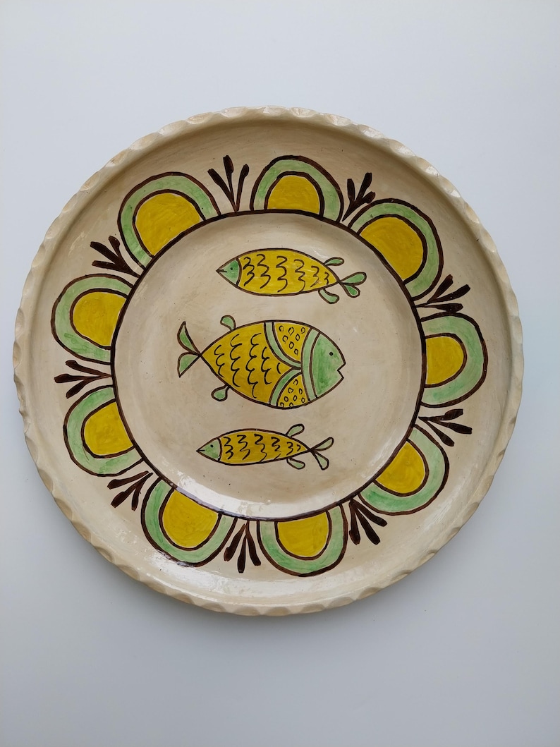 May include: A round, cream-colored ceramic plate with a scalloped edge. The plate features three yellow and green fish designs in the center. A decorative border of yellow and green arches with brown accents surrounds the fish.
